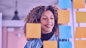 A imagem mostra uma mulher de frente para um quadro com post-its nas cores amarelo e azul. A foto sugere um processo de planejamento de go-to-market.