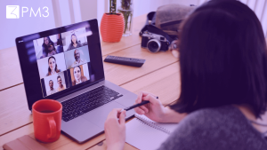 A imagem mostra uma mulher sentada em uma mesa, de frente para um notebook que exibe a tela de uma reunião de trabalho por webconferência.