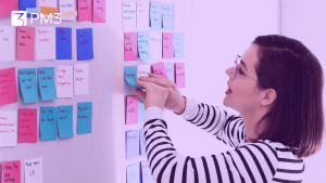 A imagem mostra uma mulher utilizando quadro com post-its para Kanban, utilizando o gerenciamento de projetos no delivery de produto,